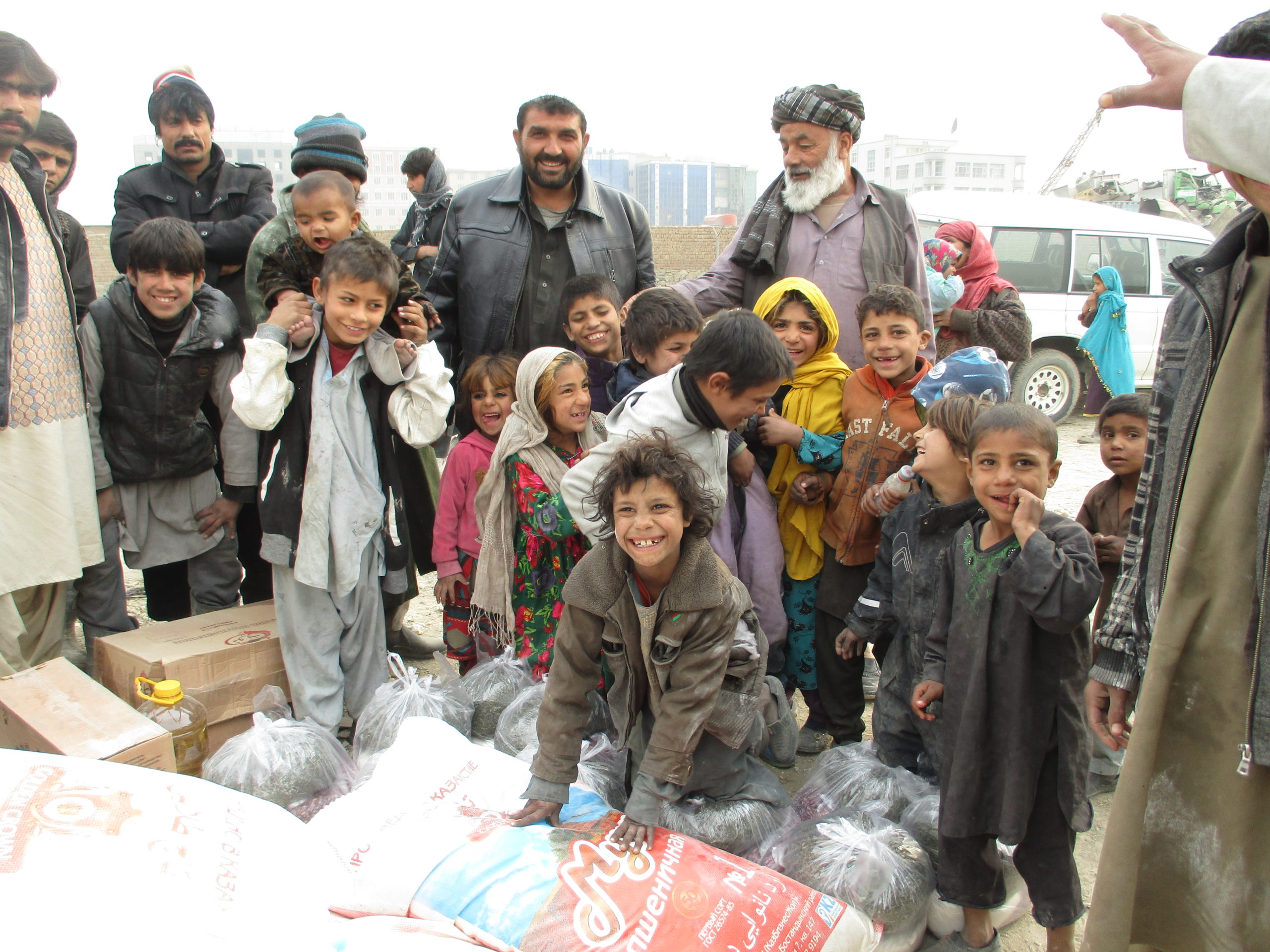 A crowd of largely happy children stand in front of a large amount of food with a few adults in the background.