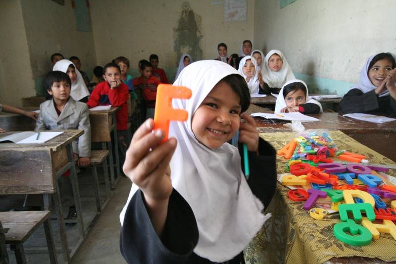 A girl with a white headscarf holds up the letter F in plastic in the foreground. To her right are more plastic letters on a table, and in the background are children sitting on wooden benches.