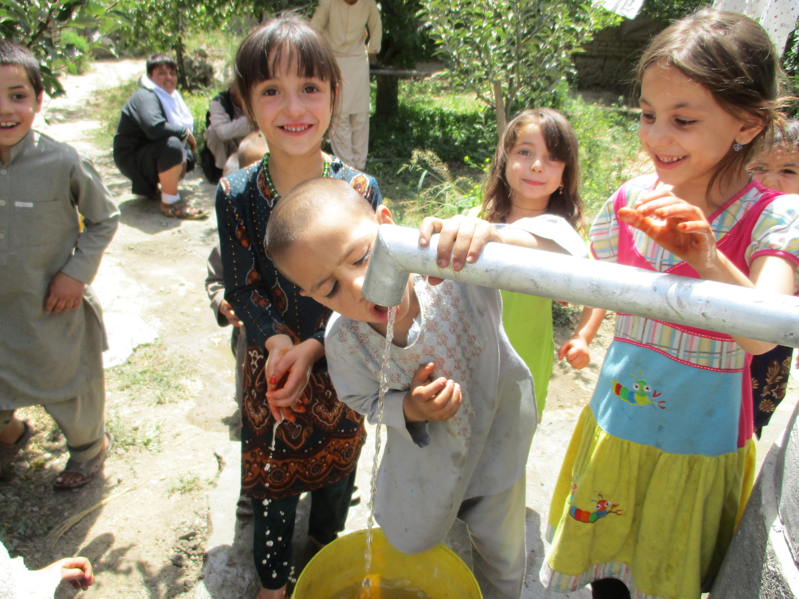 A group of children stands around a drinking fountain. A boy holds his head under the tap and drinks something. Under the tap is a yellow bucket.