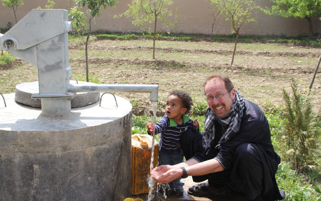 Udo Stolte kniet neben einem Trinkwasserbrunnen. Neben ihm steht ein kleiner Junge, welcher seine Hand unter den Wasserhahn hält.