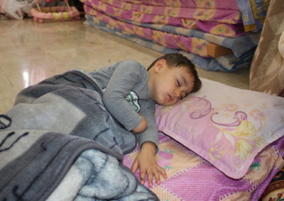 Ein Junge schläft auf ein lila-Besch karierten Decke. Er ist mit einer grauen Wolldecke zugedeckt.