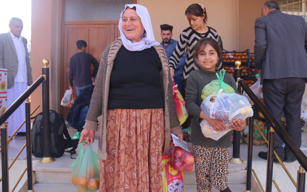 Eine ältere afghanische Frau steht mit einem Kind im Vordergrund vor eine Lebensmittelverteilung. Sie tragen Obst in Tüten und freuen sich.