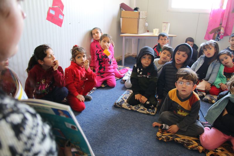 Ein Gruppe von Kindern sitzt auf dem Boden. Links im Bild steht ein Erwachsener und hält ein Buch vor seiner Brust