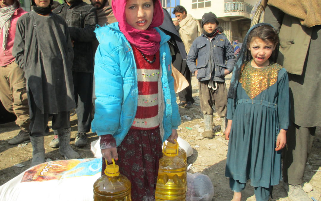 Ein Gruppe von Menschen. Im Vordergrund steht ein Mädchen mit hellblauer Jacke und rotem Kopftuch, welches drei Literpackungen mit Öl zum kochen in der Hand hält.