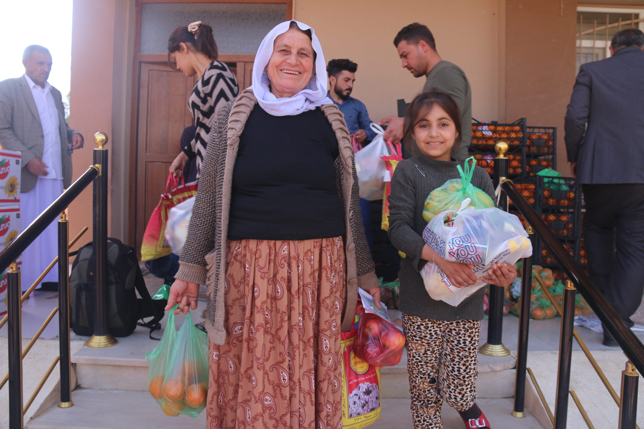 An elderly Afghan woman stands with a child in the front of a food distribution. They carry fruit in bags and are happy.